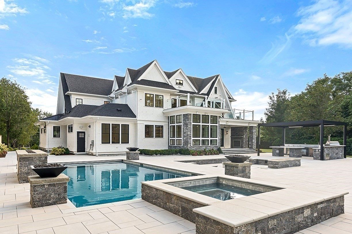 This expansive rear exterior view showcases a luxurious custom home with white horizontal siding, dark shingled roofing, and stone accents. The backyard features a large swimming pool and a raised spa, surrounded by a light-colored stone patio and several fire features. A covered outdoor kitchen area with a pergola provides a sophisticated space for entertaining, all set against a backdrop of mature trees and a clear blue sky.