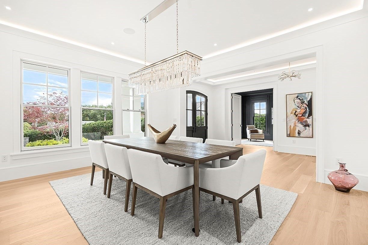 This elegant dining room features a large, rustic-style wooden table surrounded by six upholstered chairs with light-toned fabric. A striking, multi-tiered crystal chandelier hangs above the table, serving as a focal point in the bright, airy space. The room is finished with light wood flooring, a textured area rug, and crisp white walls, creating a sophisticated and welcoming atmosphere.