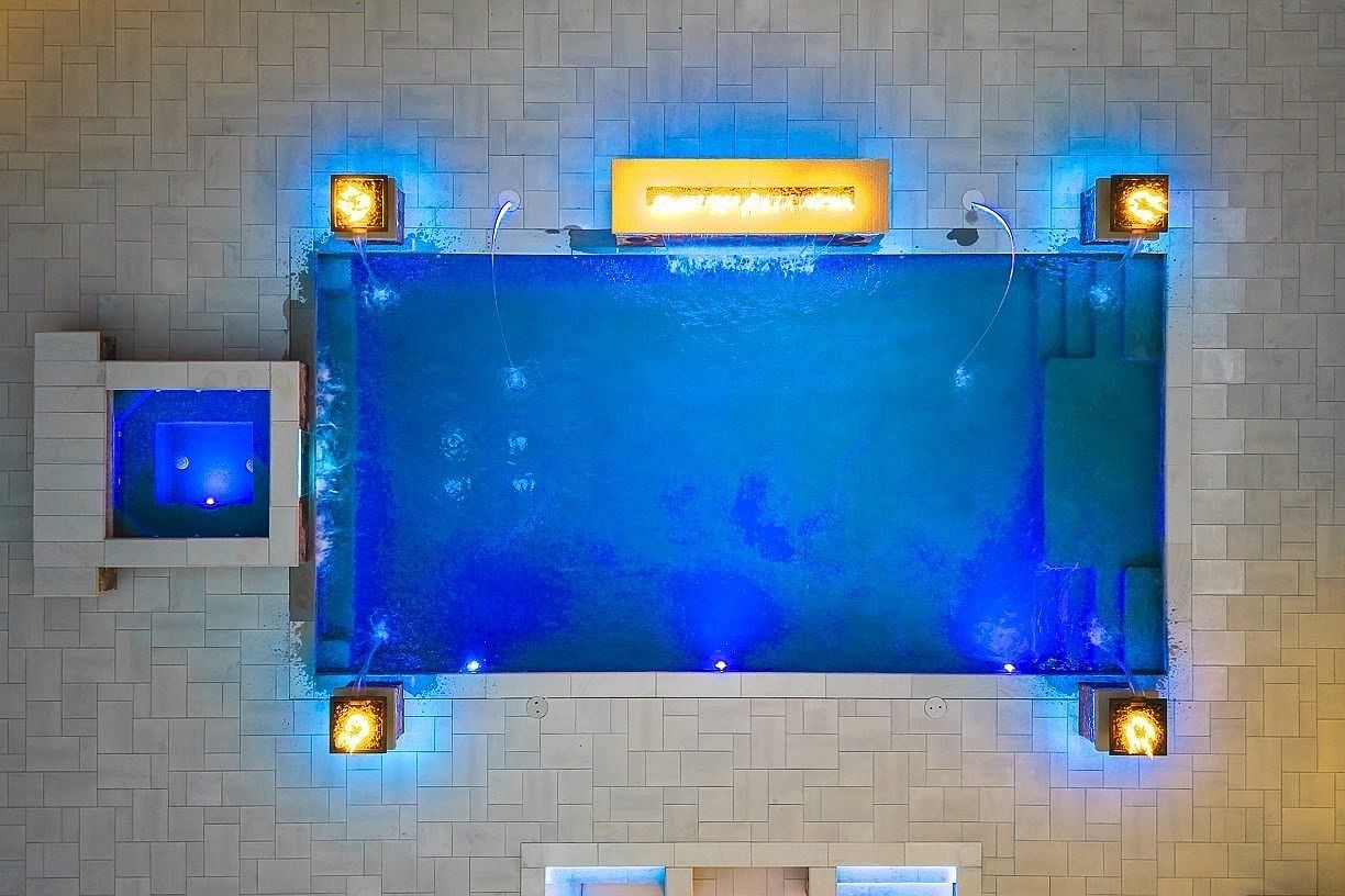 This top-down aerial view captures a luxurious rectangular swimming pool integrated with a square spa and illuminated by vibrant blue underwater lighting. The pool area is framed by four glowing fire features at the corners and a central water feature, all set within a light-toned stone patio. The composition emphasizes a symmetrical, resort-style design perfect for high-end outdoor entertaining.
