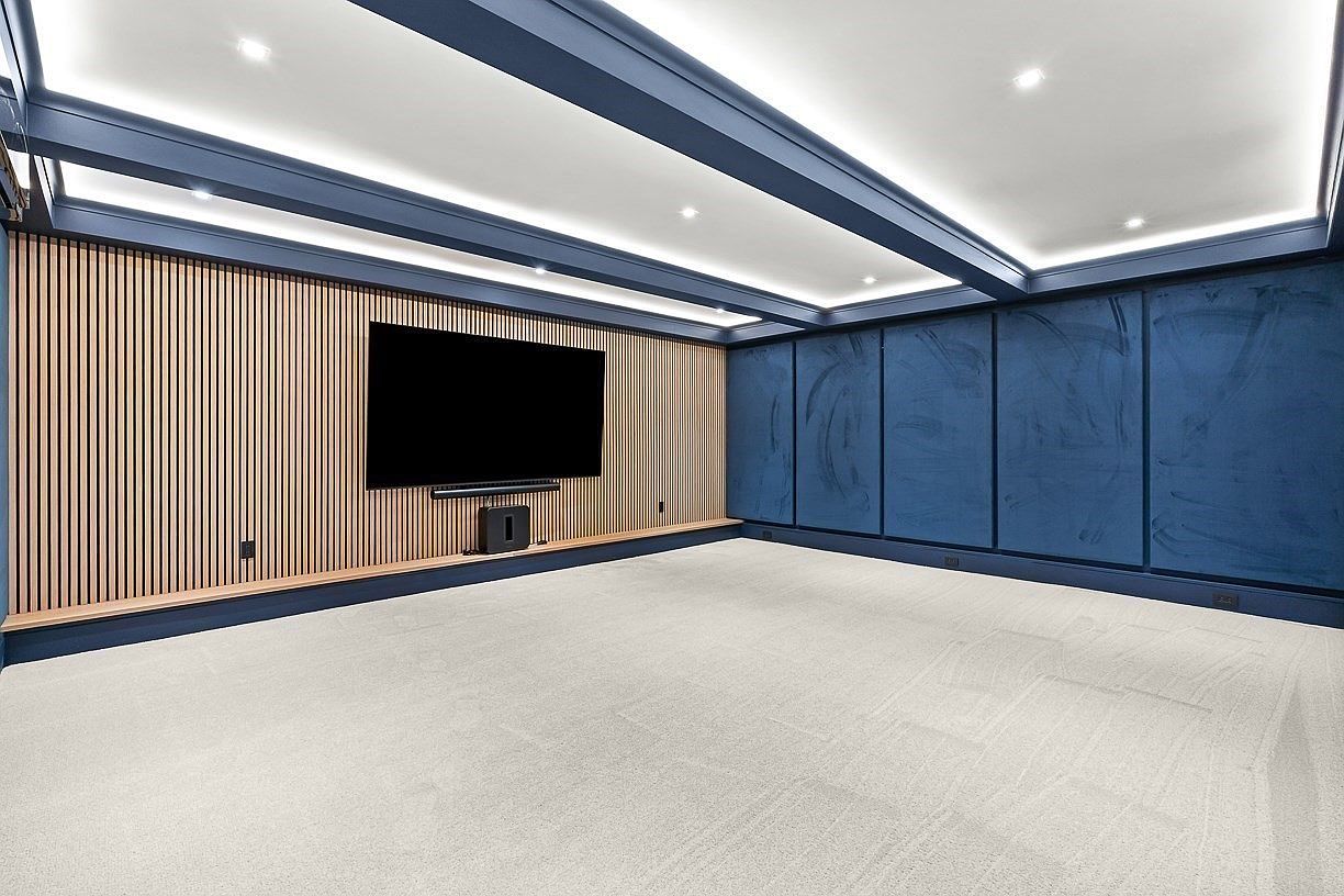 This modern home theater space features a striking accent wall with vertical light wood slats and a mounted television. The room is framed by deep blue paneled walls and a coffered ceiling with integrated recessed lighting, creating a sophisticated and immersive atmosphere. A plush, light-colored carpet covers the floor, providing a clean and comfortable foundation for this dedicated entertainment area.