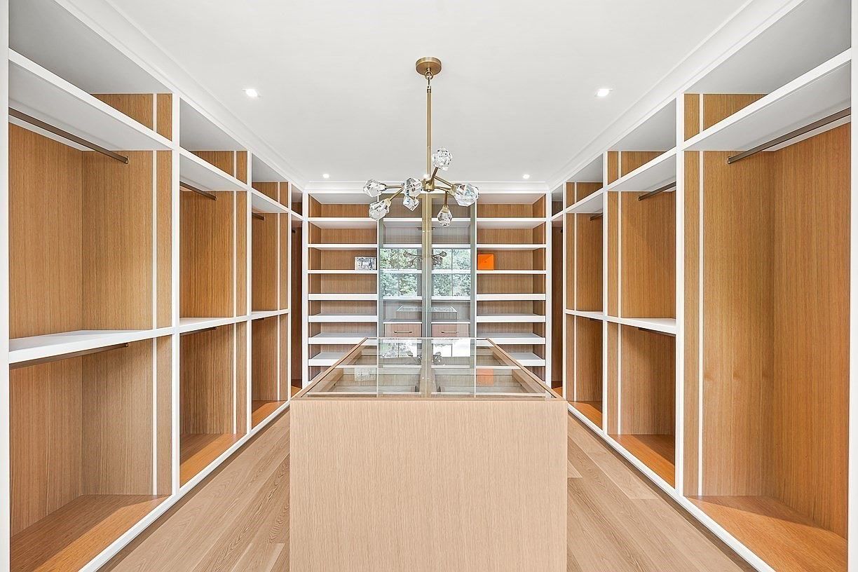 This spacious walk-in closet features extensive light wood shelving and hanging rods arranged symmetrically on both sides. A central island with a glass top provides a dedicated display area, while a modern, multi-armed chandelier hangs from the center of the ceiling. The room is bright and organized, offering a clean, contemporary aesthetic with ample storage capacity.