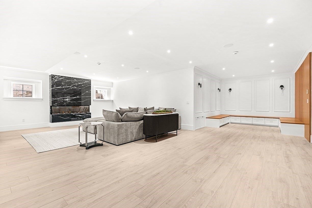 This spacious, modern living room features light-toned wood flooring and a minimalist aesthetic with crisp white walls. A prominent black stone-clad chimney breast serves as a focal point, complemented by a large grey sectional sofa and a dark console table. The room is accented by built-in white cabinetry with integrated bench seating and wall-mounted sconces, creating a clean and sophisticated atmosphere.