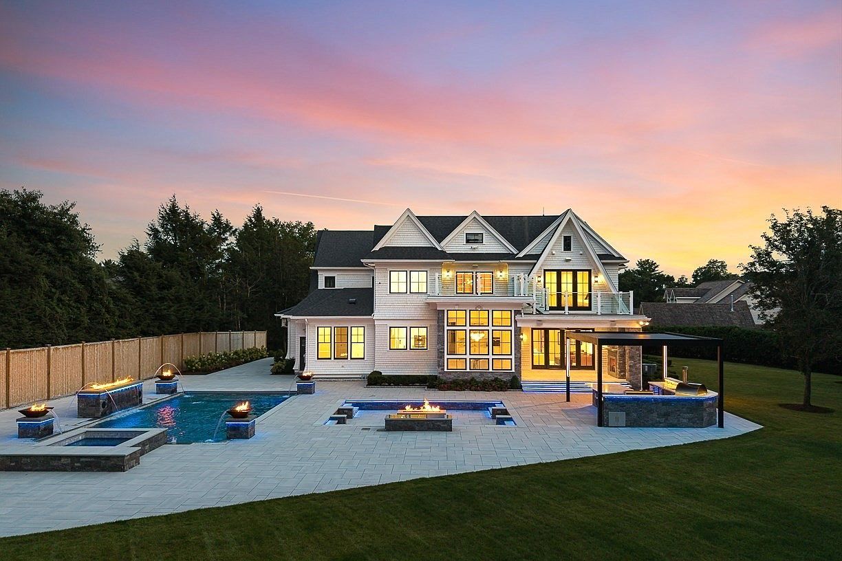 This stunning rear view of a luxury residence showcases a multi-level home with white siding and a prominent central gable. The expansive backyard features a custom swimming pool with integrated fire features, a raised spa, and a covered outdoor kitchen area under a modern pergola. The scene is captured at dusk, with the warm glow of interior and landscape lighting contrasting against a vibrant sunset sky.