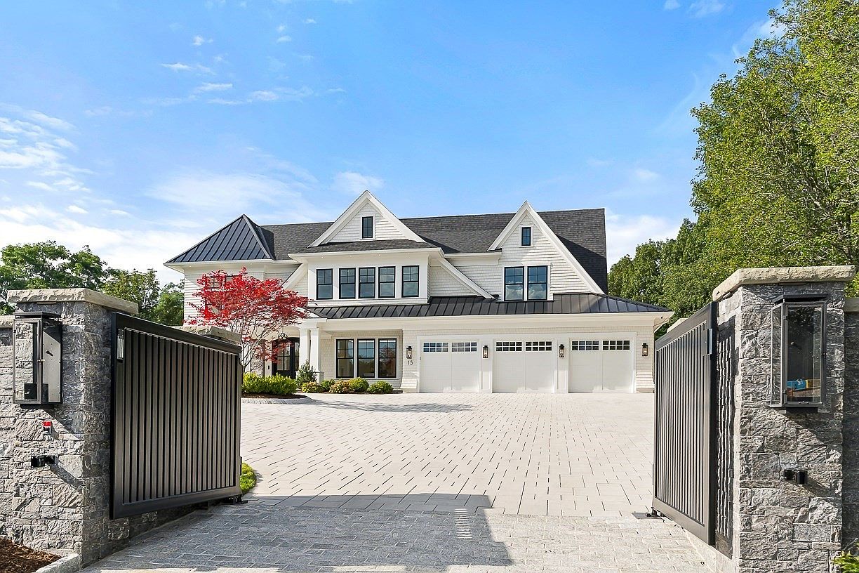 This grand, modern farmhouse-style residence is framed by a pair of imposing stone-clad gate pillars and black metal gates, creating a sense of privacy and prestige. The home features a crisp white exterior with dark metal roofing, multiple gables, and a spacious three-car garage with carriage-style doors. A wide, paved driveway leads directly to the entrance, complemented by a vibrant red-leafed tree and manicured landscaping under a bright blue sky.