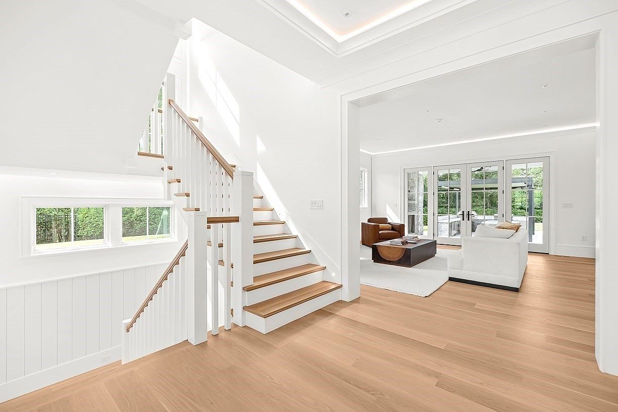 This bright and airy entryway features a staircase with light wood treads and white risers, complemented by a matching wood handrail. The open-concept design leads directly into a living area with large glass doors that provide a view of the outdoors. The space is characterized by clean white walls, light wood flooring, and a minimalist aesthetic that creates a welcoming and spacious atmosphere.