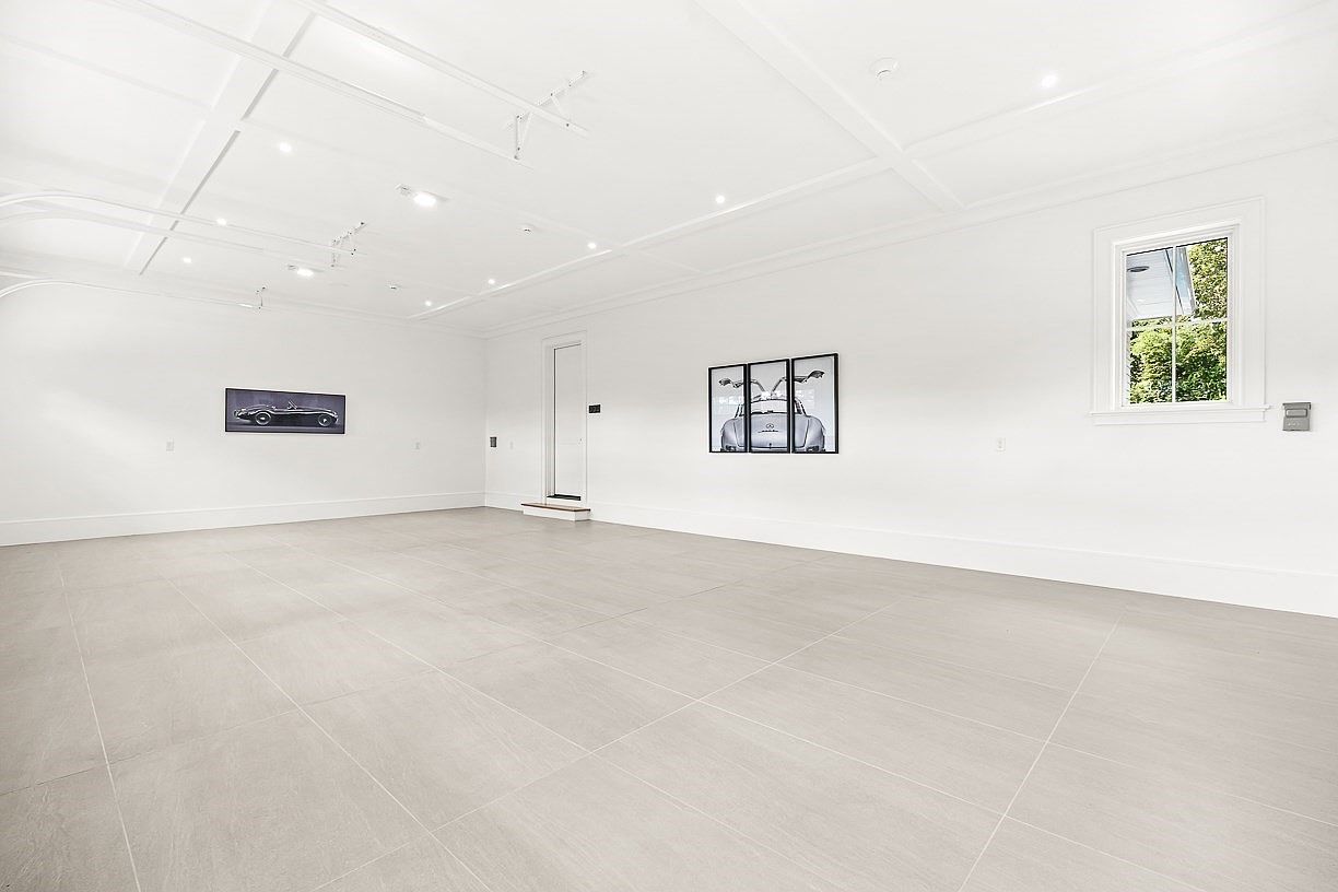 This spacious, minimalist garage features a clean, white aesthetic with large-format light-toned floor tiles and a coffered ceiling. The room is sparsely decorated with two framed automotive-themed art pieces on the walls and a single window that allows natural light to enter. The perspective is wide and centered, emphasizing the expansive, uncluttered floor space suitable for vehicle storage or a multi-purpose area.