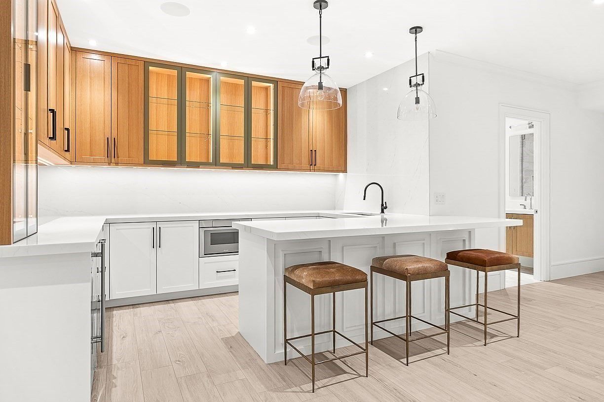 This bright, contemporary kitchen features a clean aesthetic with white lower cabinetry and light wood upper cabinets, including glass-fronted display units. A large central island with a stone countertop provides seating for three, complemented by modern pendant lighting and a sleek faucet. The space is finished with light-toned flooring and a minimalist backsplash, creating an airy and sophisticated atmosphere.