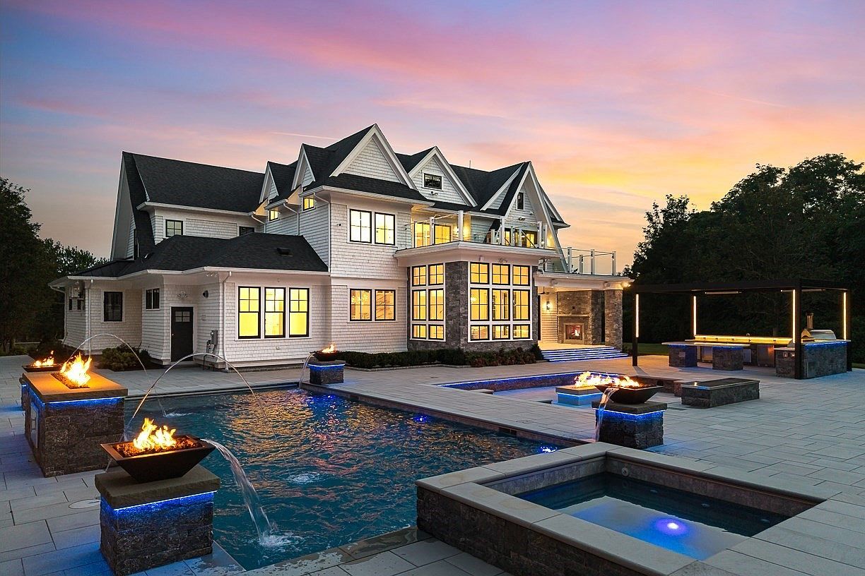 This stunning rear exterior view showcases a luxurious multi-story home with white siding and dark roofing, set against a vibrant sunset sky. The expansive backyard features a sophisticated pool and spa complex with integrated fire features, stone-clad water bowls, and a covered outdoor kitchen area. The scene is captured from a slightly elevated perspective, emphasizing the seamless transition between the elegant architecture and the high-end resort-style outdoor living space.
