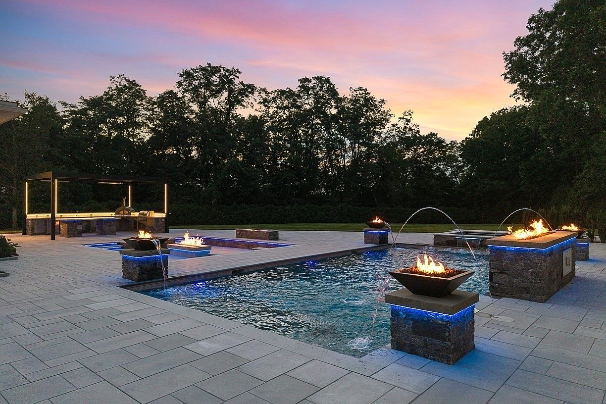 This luxurious outdoor living space features a custom swimming pool accented by multiple fire bowls and integrated water features that create a dramatic evening ambiance. A paved patio surrounds the pool, leading to a covered outdoor kitchen area equipped with a pizza oven and modern lighting. The scene is captured at dusk, highlighting the vibrant sunset sky and the glowing blue LED lighting integrated into the pool and stone structures.