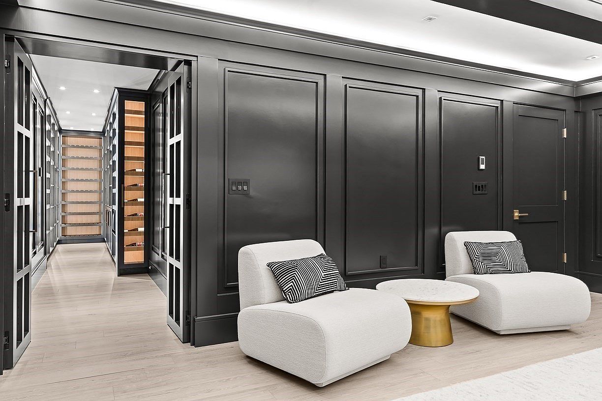 This sophisticated lounge area features dark, paneled walls and two plush, cream-colored armchairs accented with geometric-patterned throw pillows. A small, round table with a stone top and a metallic base sits between the chairs, while an open doorway leads into a climate-controlled wine cellar with extensive bottle storage. The space is finished with light wood flooring, creating a refined and modern aesthetic perfect for entertaining.