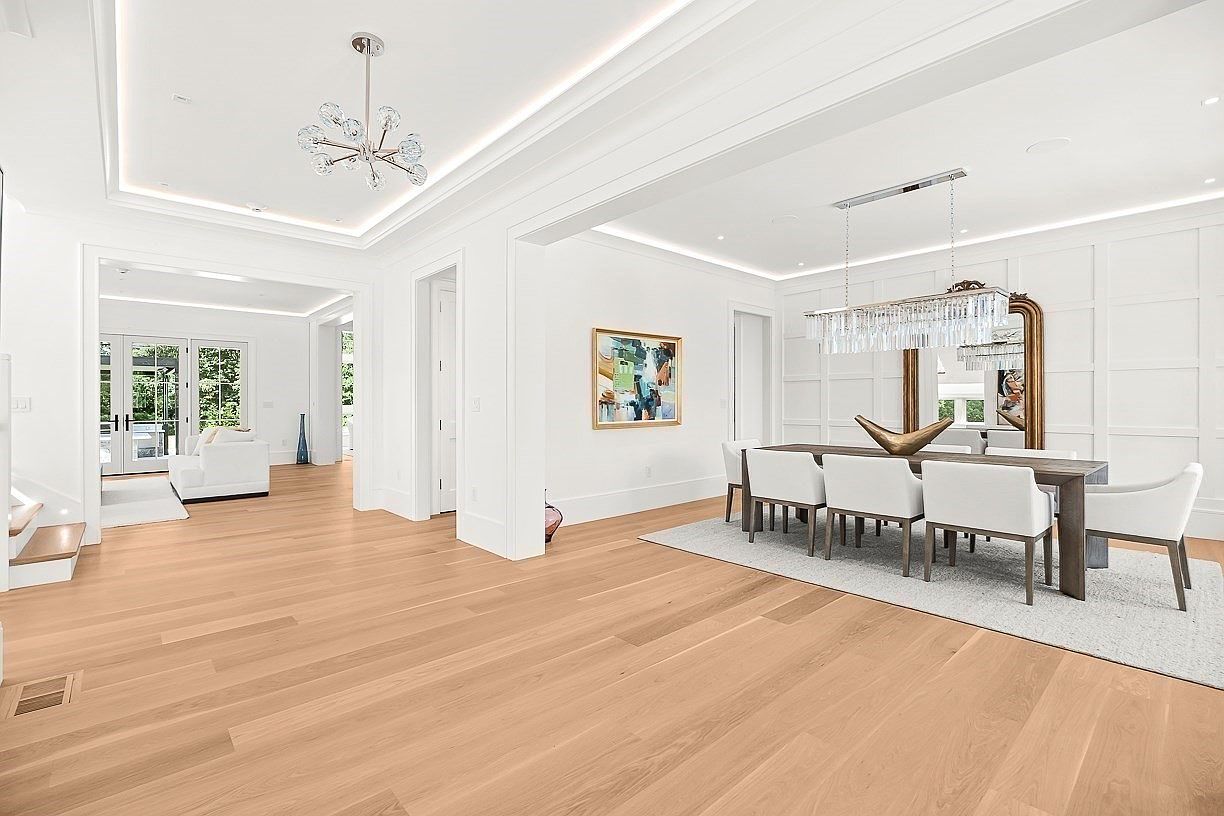 This bright and airy open-concept space features a formal dining area on the right, anchored by a long table with white upholstered chairs and a large, modern crystal chandelier. The room showcases light wood flooring, crisp white walls, and elegant crown molding, with a view into a secondary living area through a wide transition. A large, decorative anchor sculpture sits as a centerpiece on the dining table, adding a unique nautical touch to the sophisticated, minimalist interior.