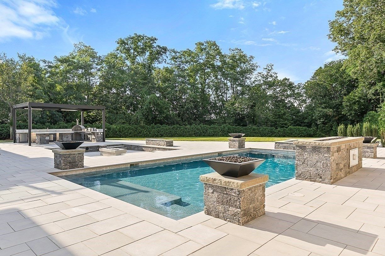 This luxurious outdoor living space features a rectangular swimming pool with a built-in tanning ledge, surrounded by light-toned stone pavers. The area is accented by multiple stone-clad fire bowls and a covered outdoor kitchen structure equipped with a pizza oven and grill. The scene is framed by a lush backdrop of mature trees, creating a private and sophisticated resort-style atmosphere.