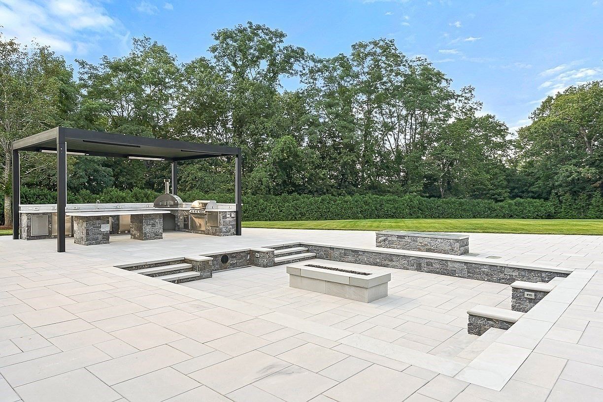This expansive outdoor living space features a sunken lounge area with a central stone fire pit and tiered seating walls. To the left, a covered outdoor kitchen is equipped with a pizza oven, stainless steel grill, and stone-clad counters under a modern pergola. The area is paved with large, light-toned stone tiles and overlooks a manicured lawn bordered by a dense hedge and mature trees.