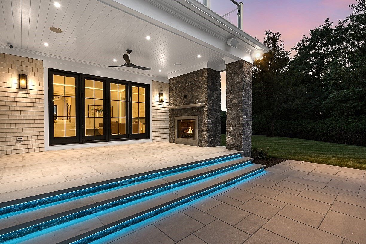 This sophisticated covered patio features large glass doors that open to a spacious stone-paved terrace. A prominent stone-clad chimney breast houses an outdoor fireplace, while the steps leading down to the yard are accented with modern blue LED strip lighting. The space is finished with a clean white ceiling, recessed lighting, and a ceiling fan, creating an inviting atmosphere for evening relaxation.