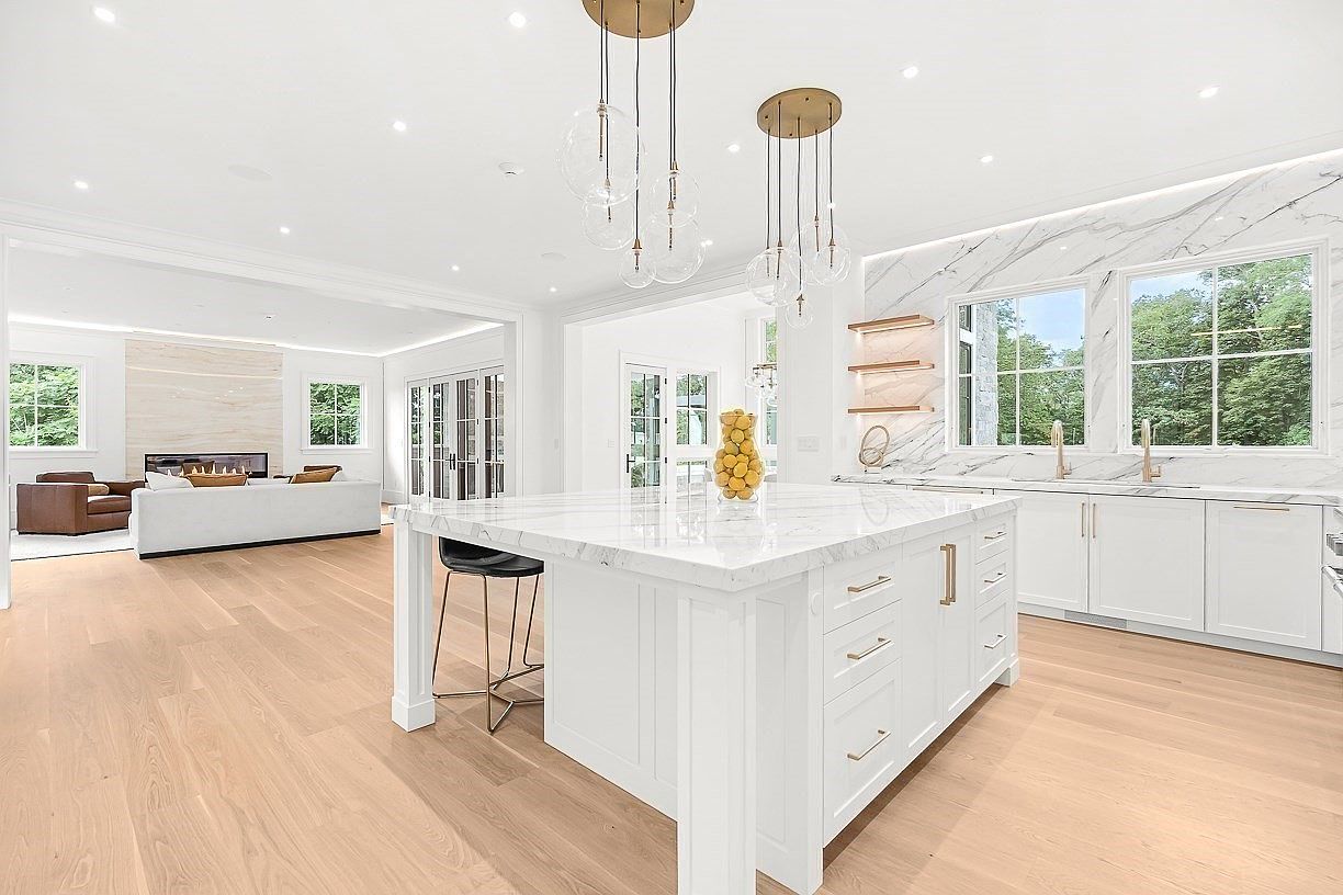 This bright, open-concept kitchen features a large central island with a stone countertop and integrated seating, serving as the focal point of the space. White cabinetry and light wood flooring create a clean, airy aesthetic, while modern pendant lights hang above the island. The perspective looks from the kitchen toward an adjacent living area, which includes a sofa and a prominent stone-clad chimney breast, emphasizing the home's spacious and cohesive design.