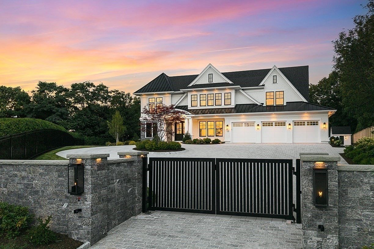 This striking front view captures a luxurious modern farmhouse residence at dusk, framed by a stone-walled gated entrance. The home features a crisp white exterior with dark metal roofing, multiple gables, and a spacious three-car garage, all set against a vibrant, colorful sunset sky. A paved driveway leads through the gate toward the grand entrance, emphasizing the property's privacy and upscale curb appeal.