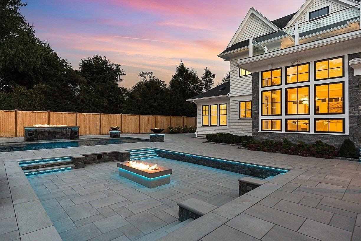 This luxurious backyard features a sunken fire pit area with integrated blue LED lighting, surrounded by a spacious stone-paved patio. A swimming pool with a water feature sits adjacent to the seating area, all set against the backdrop of a modern home with large windows and a sunset sky. The scene is captured from a low angle, emphasizing the sophisticated outdoor living space and high-end landscaping.