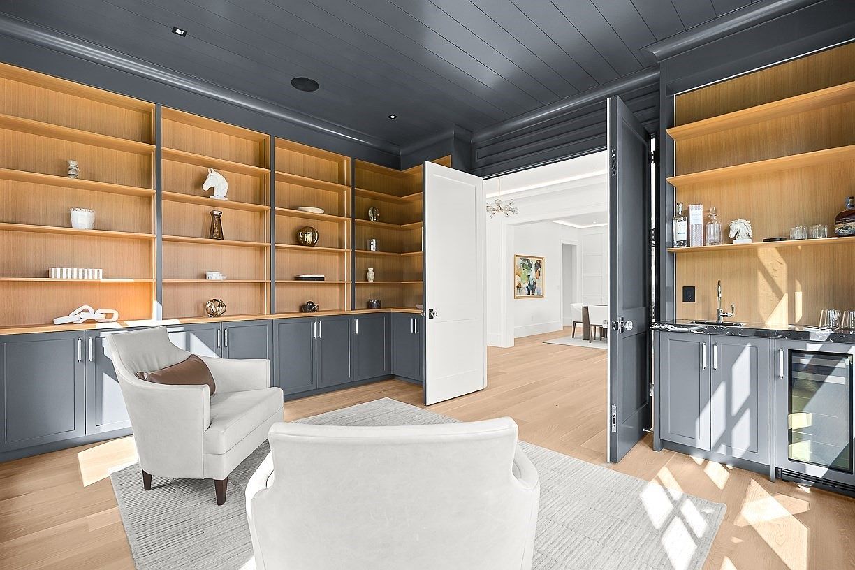 This sophisticated home office or study features custom built-in shelving with light wood interiors contrasted against dark cabinetry. Two plush, light-colored armchairs are arranged on a textured area rug, creating a comfortable seating area, while a wet bar with a beverage refrigerator is integrated into the cabinetry on the right. The room is finished with a dark-painted ceiling and light wood flooring, with an open doorway leading into a bright, adjacent living space.