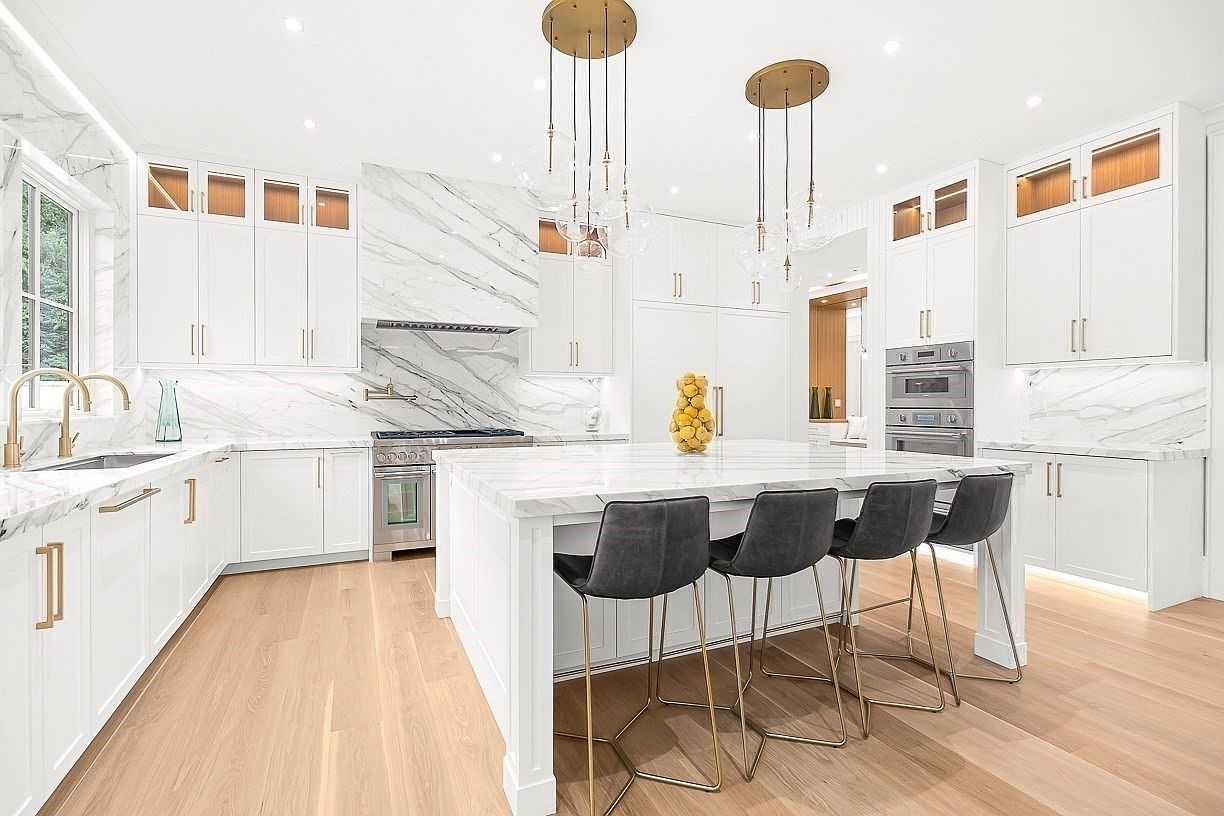 This bright, modern kitchen features white cabinetry, a large central island with seating for four, and light-toned wood flooring. The space is accented by a striking stone backsplash and matching countertops, complemented by gold-toned hardware and elegant pendant lighting. The perspective is a wide-angle shot from the kitchen floor, showcasing the clean lines and high-end finishes of the open-concept layout.