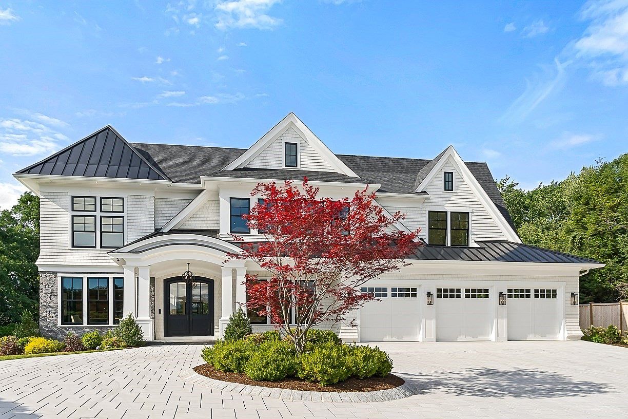 This impressive two-story luxury residence features a sophisticated blend of white siding, stone accents, and a dark metal roof. The front facade is highlighted by a grand arched entryway, a circular driveway with a central landscaped island, and a three-car garage. A vibrant red Japanese maple tree serves as a focal point in the front yard, enhancing the home's curb appeal under a bright blue sky.