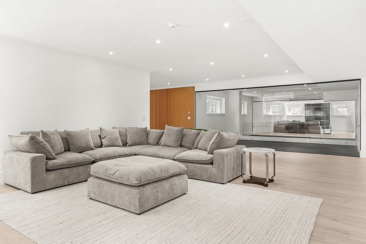 This spacious living area features a large, light-colored sectional sofa paired with a matching oversized ottoman, centered on a textured area rug. The room is characterized by a minimalist aesthetic with light wood flooring and white walls, complemented by a striking glass-walled partition that reveals an adjacent room. The perspective is eye-level, capturing the open-concept layout and the clean, contemporary design of the space.