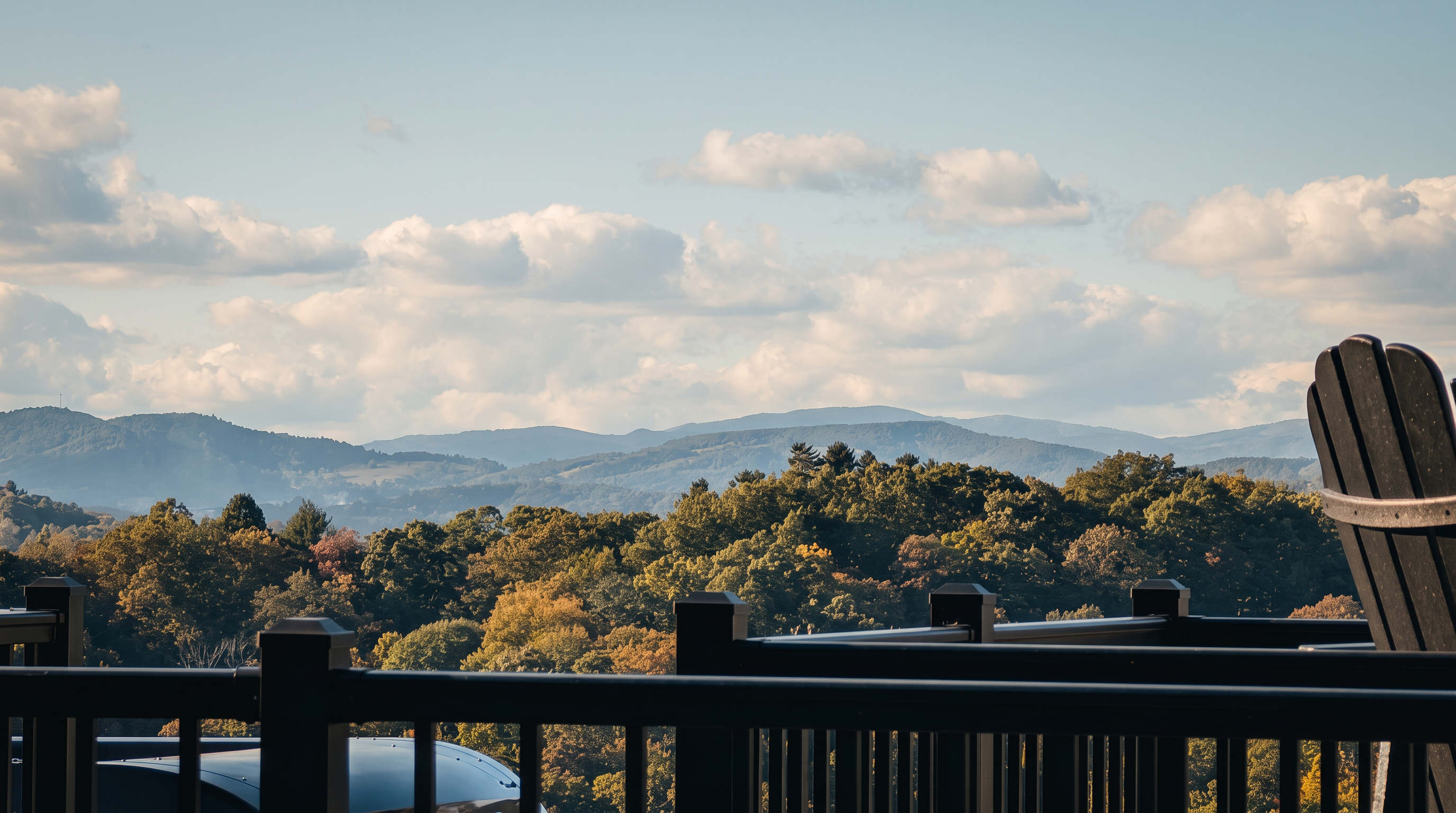 This image captures a scenic view from an elevated deck, featuring a black railing that frames a vast, rolling mountain landscape. The foreground includes a portion of a dark-colored outdoor chair, emphasizing the relaxing nature of the space. The expansive sky filled with soft, scattered clouds complements the lush, tree-covered hills in the distance, creating a serene and picturesque atmosphere.