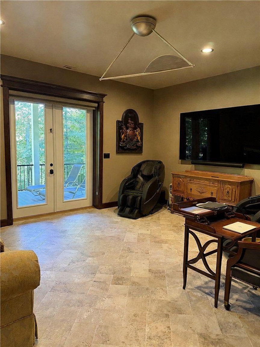 This interior space features light-colored stone tile flooring and a neutral wall palette, creating a versatile room currently set up as a home office and relaxation area. A prominent black massage chair sits near a wall-mounted crest, while a wooden desk and a media console with a large television provide functional workspace and entertainment. Double glass doors lead out to a deck, allowing natural light to fill the room and offering a view of the surrounding trees.