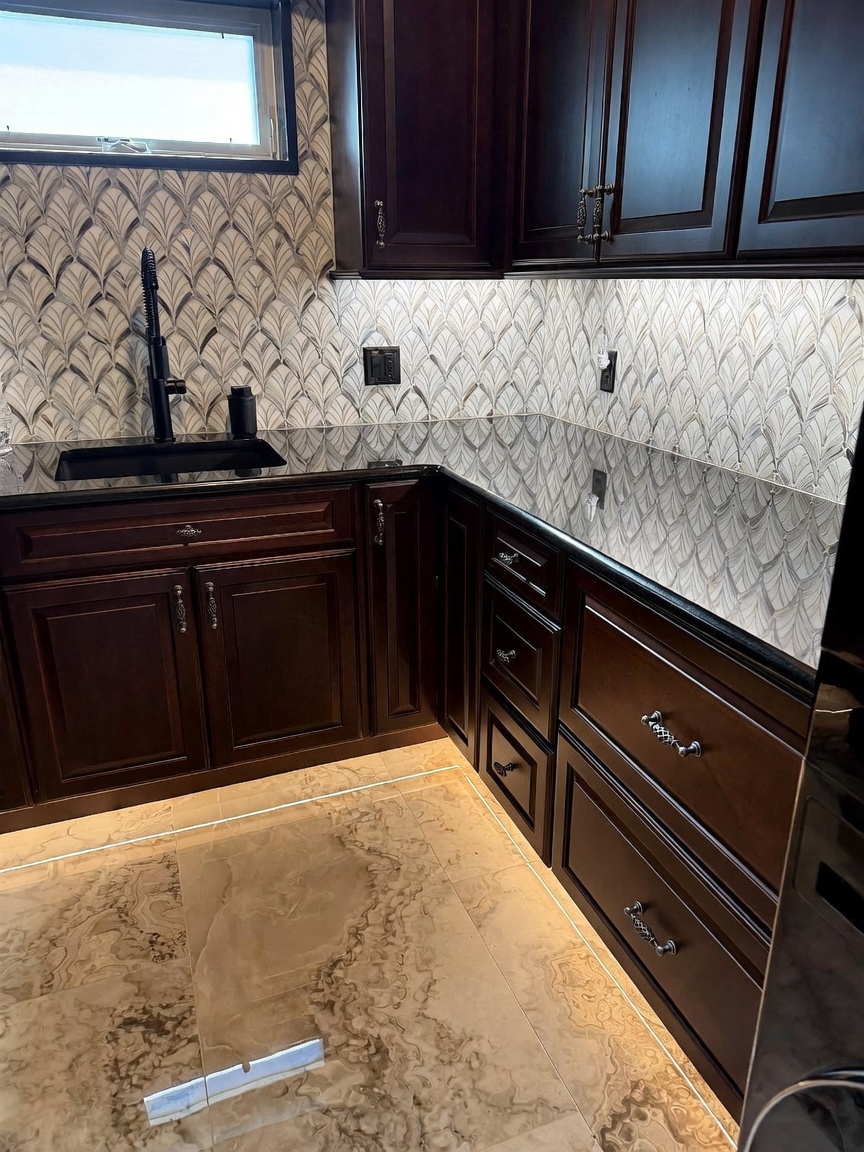 This kitchen area features dark, rich-toned cabinetry paired with a polished black countertop and a distinctive, patterned tile backsplash. A modern black faucet is mounted over an undermount sink, while under-cabinet lighting highlights the workspace and the textured stone flooring. The perspective is a close-up, eye-level shot that emphasizes the high-contrast color palette and the sleek, contemporary design of the cabinetry hardware.