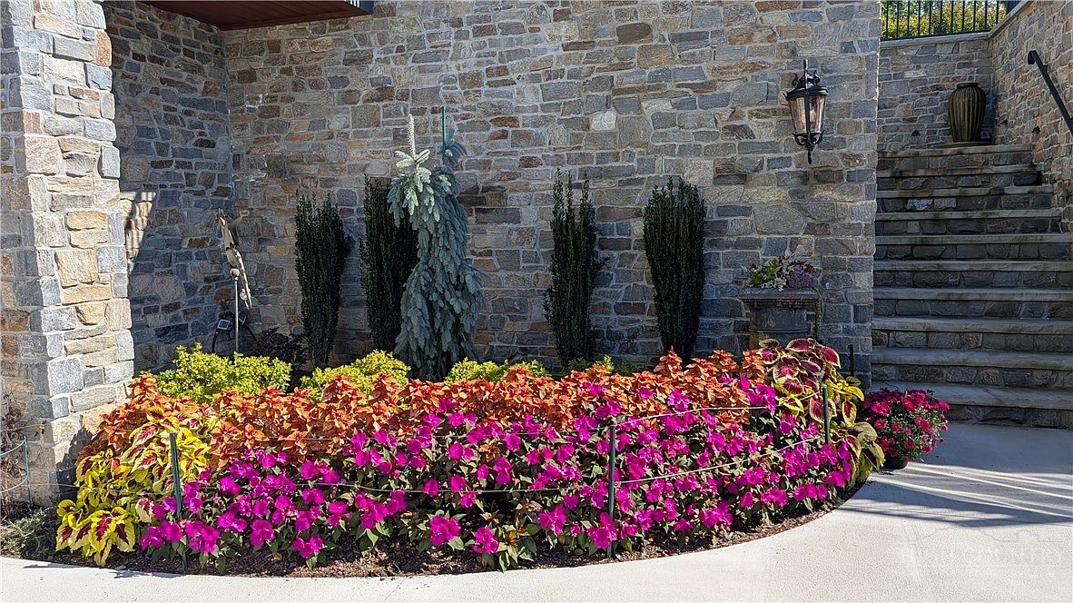 This exterior view showcases a meticulously landscaped garden bed set against a rustic stone wall. The garden features a vibrant mix of bright pink flowers, orange-toned foliage, and tall, slender evergreens, all bordered by a low wire fence. To the right, a stone staircase leads upward, framed by the textured masonry of the home's facade and a classic wall-mounted lantern.