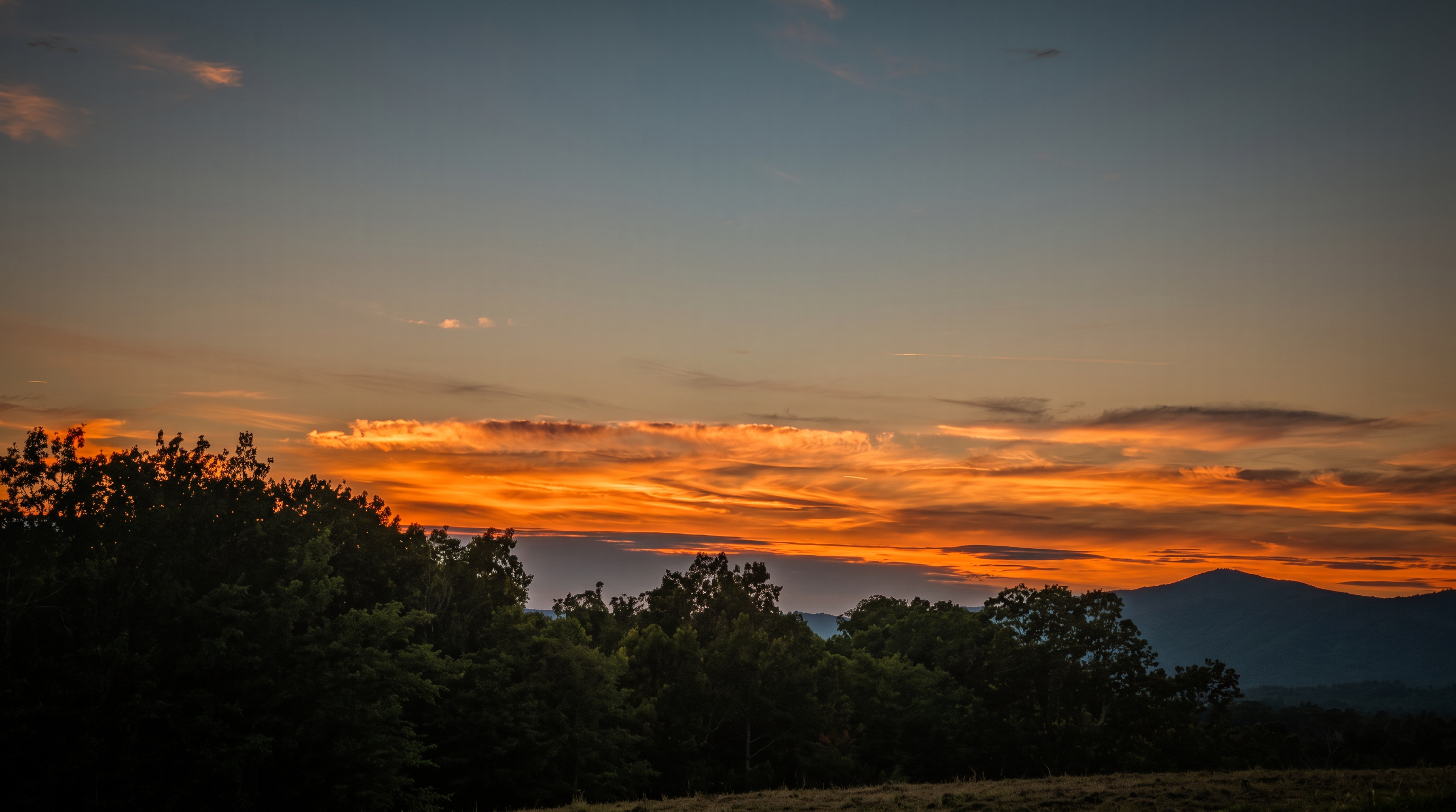 This image captures a dramatic sunset view over a mountainous landscape, showcasing a vibrant sky filled with shades of orange, red, and deep blue. In the foreground, the dark silhouettes of trees frame the horizon, while the rolling outlines of distant mountains are visible against the glowing sky. The perspective is a wide, scenic shot that emphasizes the natural beauty and tranquil atmosphere of the property's surroundings.