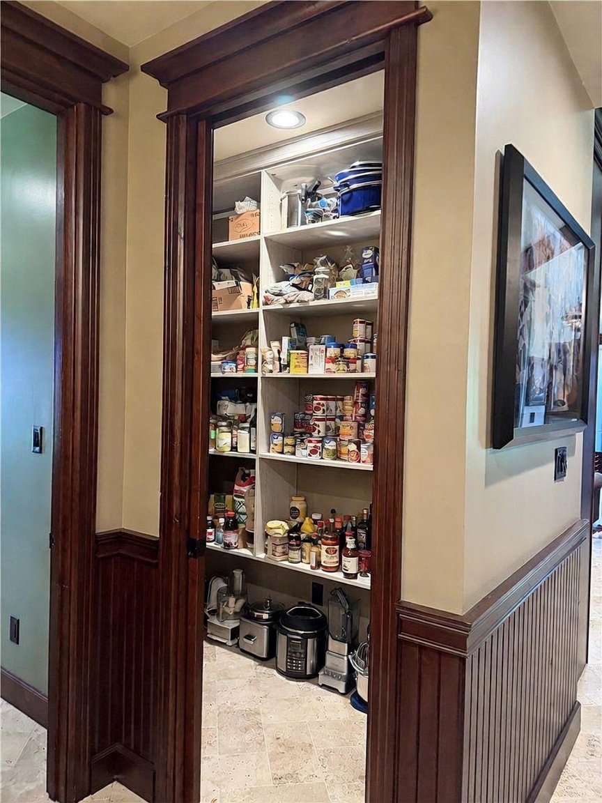 This walk-in pantry features custom built-in shelving with a dark wood trim frame that matches the surrounding wainscoting. The interior is organized with various food items, canned goods, and small kitchen appliances stored on the lower level. The space is illuminated by a recessed ceiling light, creating a functional and well-organized storage area.