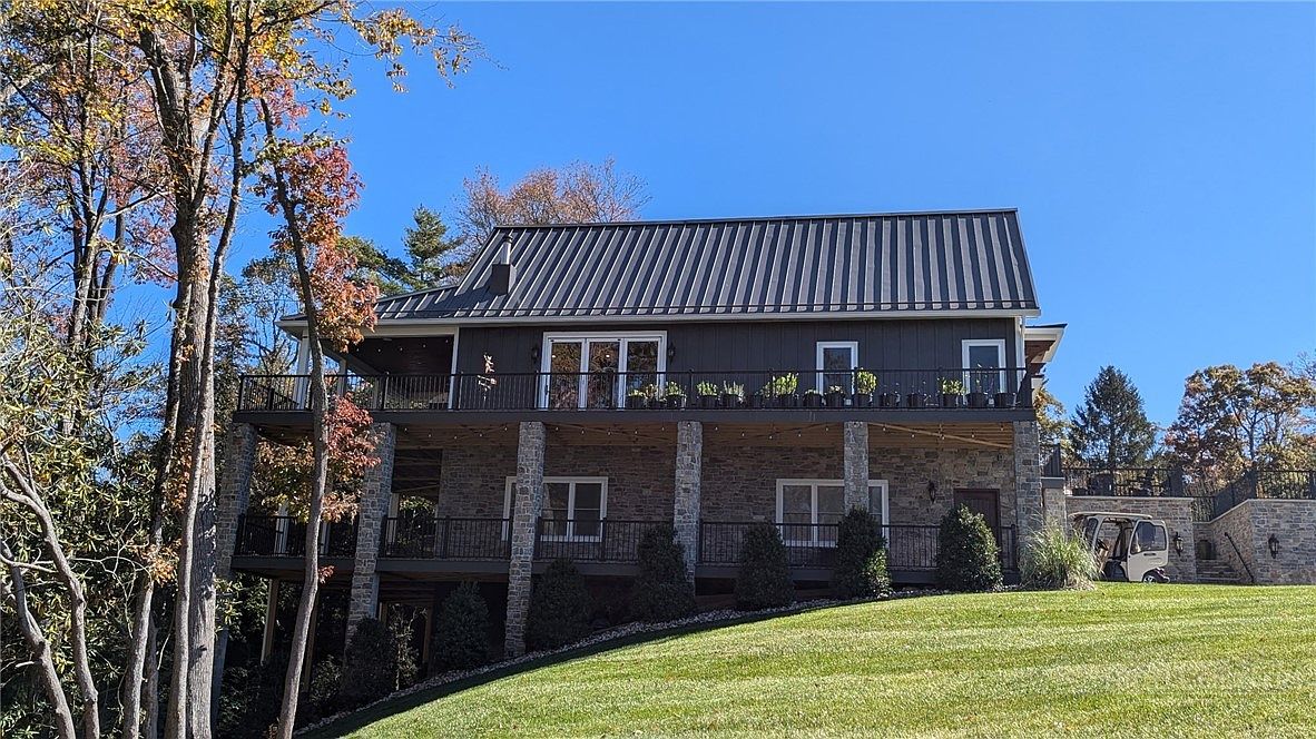 This rear view of a multi-level home showcases a blend of dark vertical siding and natural stone masonry. The property features a spacious upper deck and a lower-level patio, both enclosed by black metal railings, overlooking a manicured lawn. The structure is topped with a metal roof and is surrounded by mature trees under a clear blue sky.
