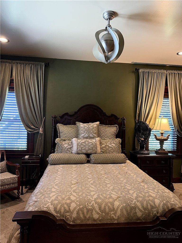 This primary bedroom features a dark wood bed frame centered against a deep olive-green wall, flanked by two windows dressed in neutral-toned curtains. The bed is adorned with a patterned quilt and a collection of decorative pillows, while a bedside table with a lamp and a small fan sits to the right. The room is illuminated by a unique, modern spherical ceiling fixture, creating a cozy and traditional atmosphere.