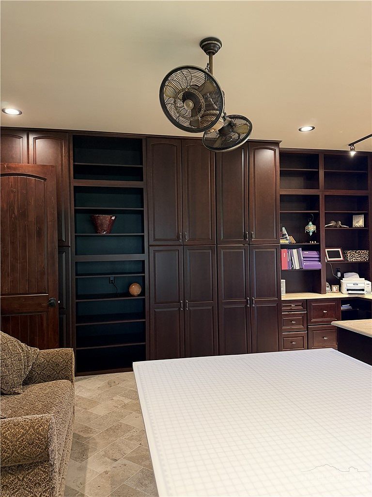 This home office or craft room features extensive dark wood cabinetry and built-in shelving that provides ample storage and display space. A large, grid-patterned work table occupies the foreground, while a comfortable armchair sits to the side, creating a functional and organized workspace. The room is illuminated by recessed lighting and a unique dual-fan ceiling fixture, with stone-look tile flooring completing the professional aesthetic.