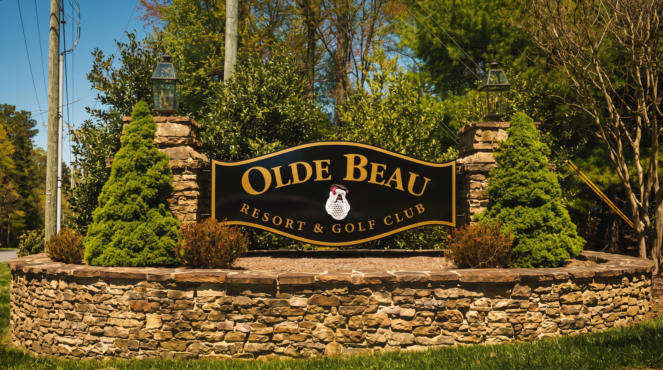 This image features the prominent entrance sign for Olde Beau Resort & Golf Club, set against a backdrop of lush green trees and a clear blue sky. The sign is elegantly mounted on a curved stone wall with symmetrical landscaping, including manicured evergreen shrubs and decorative pillar lamps. This welcoming display highlights the well-maintained and upscale nature of the resort community.