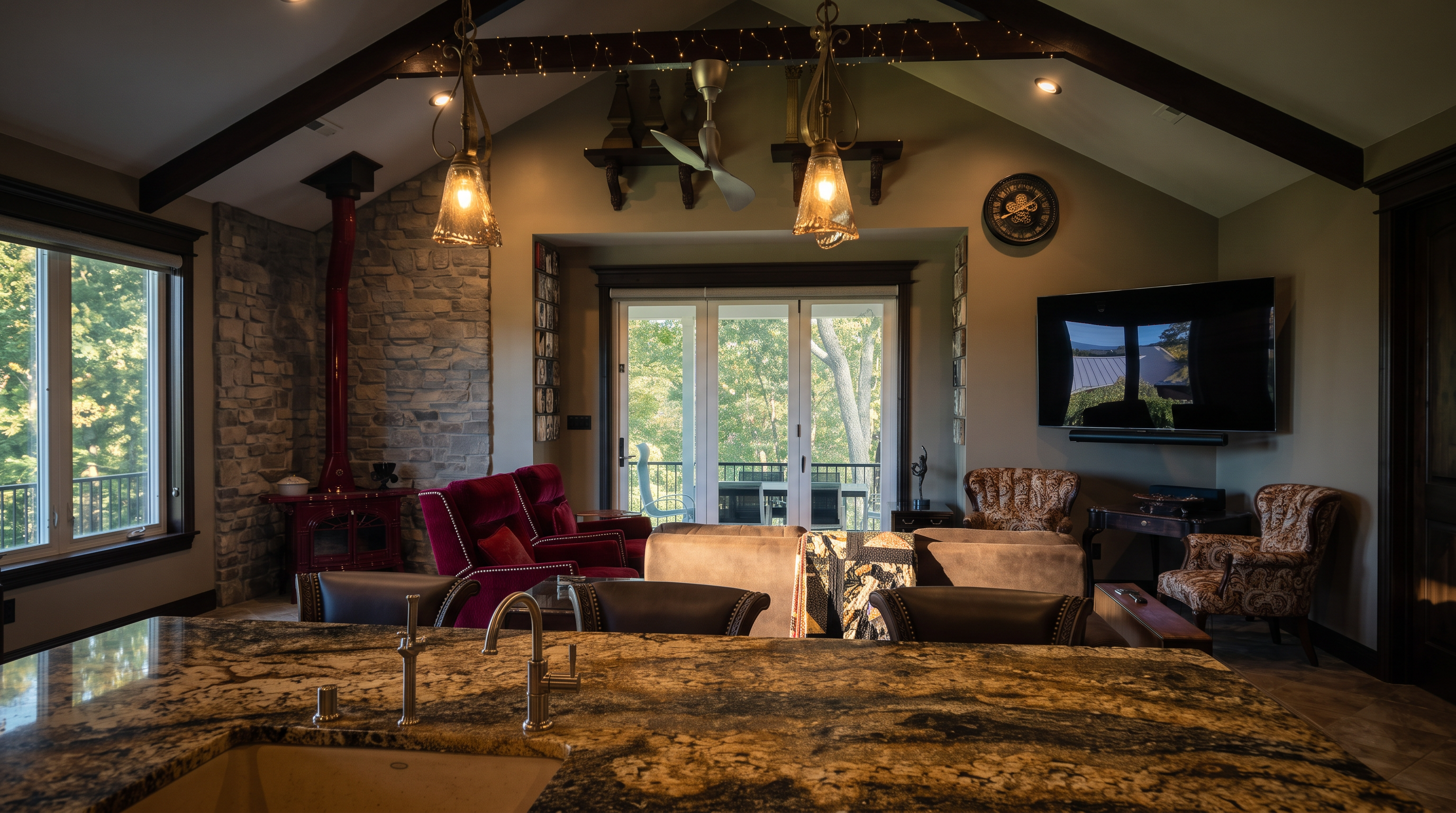 This open-concept living space features a vaulted ceiling with exposed dark wood beams and decorative string lights. A prominent stone-clad chimney breast anchors the left side of the room, while a large granite-topped island in the foreground provides a transition from the kitchen area. The room is furnished with plush seating, including deep red armchairs and a dark sofa, all oriented toward a wall-mounted television and a set of glass doors that open to an outdoor deck.