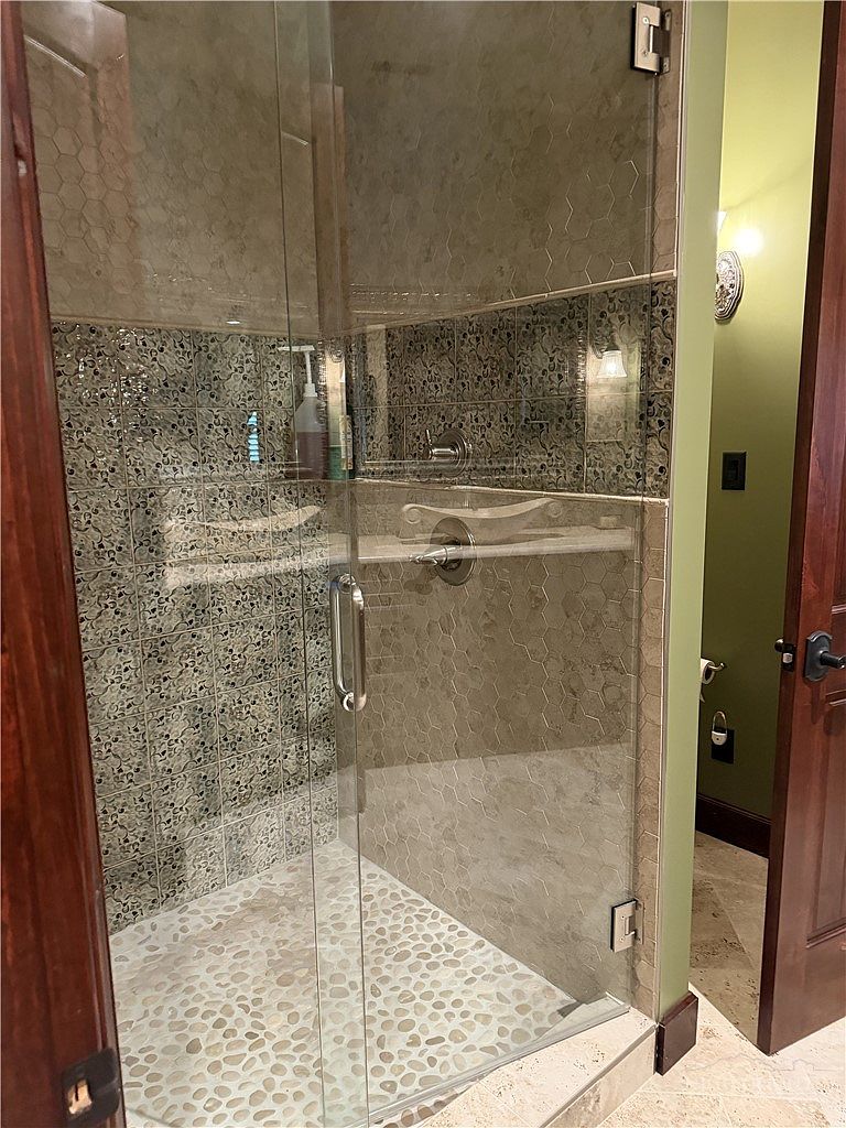 This bathroom features a glass-enclosed walk-in shower with a pebble-textured floor and decorative patterned wall tiles. A built-in stone shelf provides storage within the shower, while the adjacent wall is painted a soft green. The perspective captures the shower entrance from a hallway, highlighting the transition between the tiled shower area and the surrounding room.