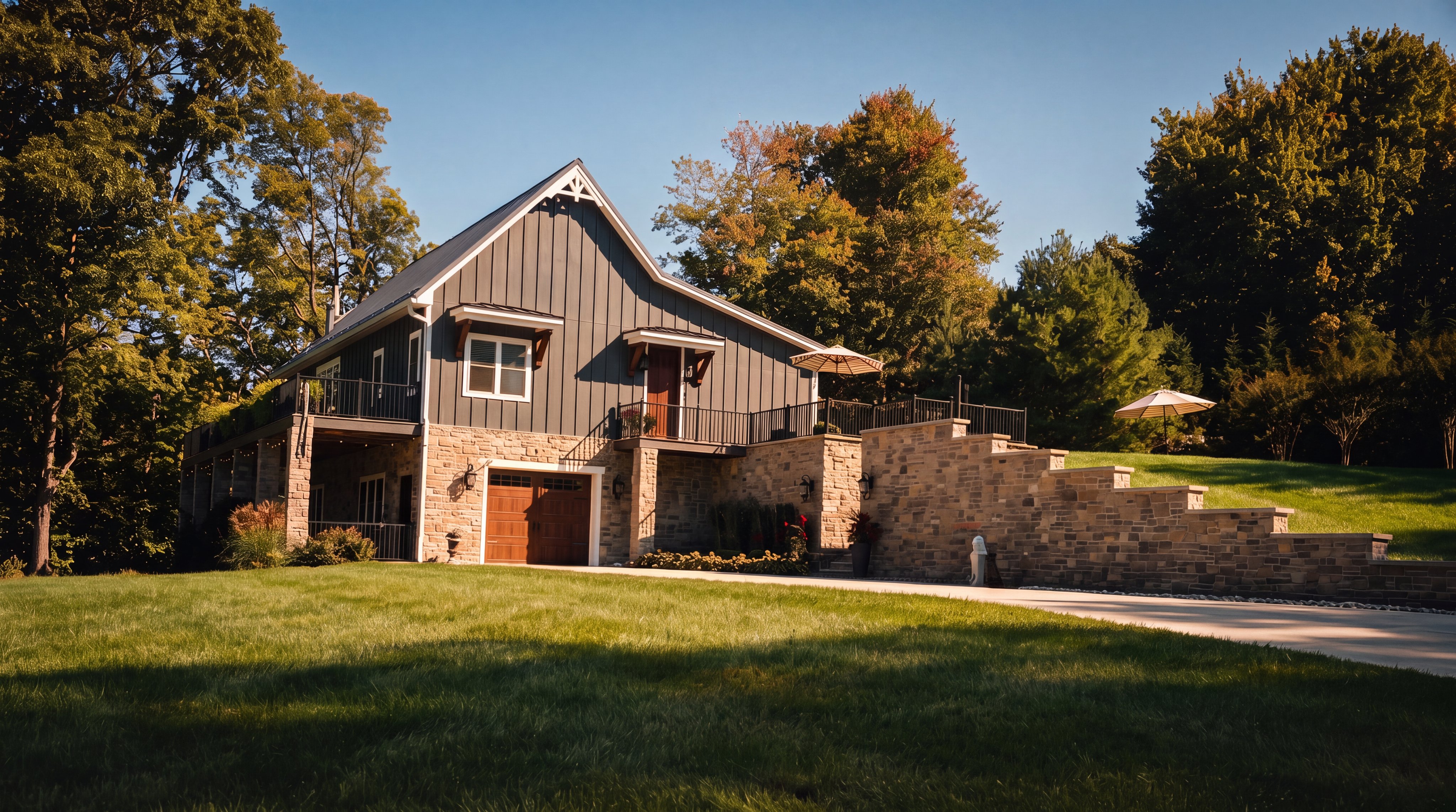 This striking home features a modern farmhouse aesthetic with dark vertical siding contrasted by a substantial stone-clad lower level. A paved driveway leads to a garage entrance, while a stone retaining wall defines the terraced landscaping and leads toward an elevated deck area. The property is set against a backdrop of mature trees under a clear blue sky, presenting a well-maintained and inviting curb appeal.