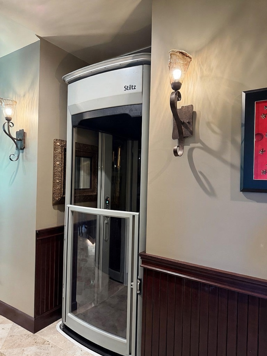 This image captures a Stiltz residential elevator installed within a home hallway, featuring a sleek, curved glass and metal design. The elevator is flanked by decorative wall sconces with textured glass shades, set against neutral-toned walls with dark wood wainscoting. The perspective is a straight-on, eye-level shot that highlights the integration of modern accessibility features into a traditional interior aesthetic.