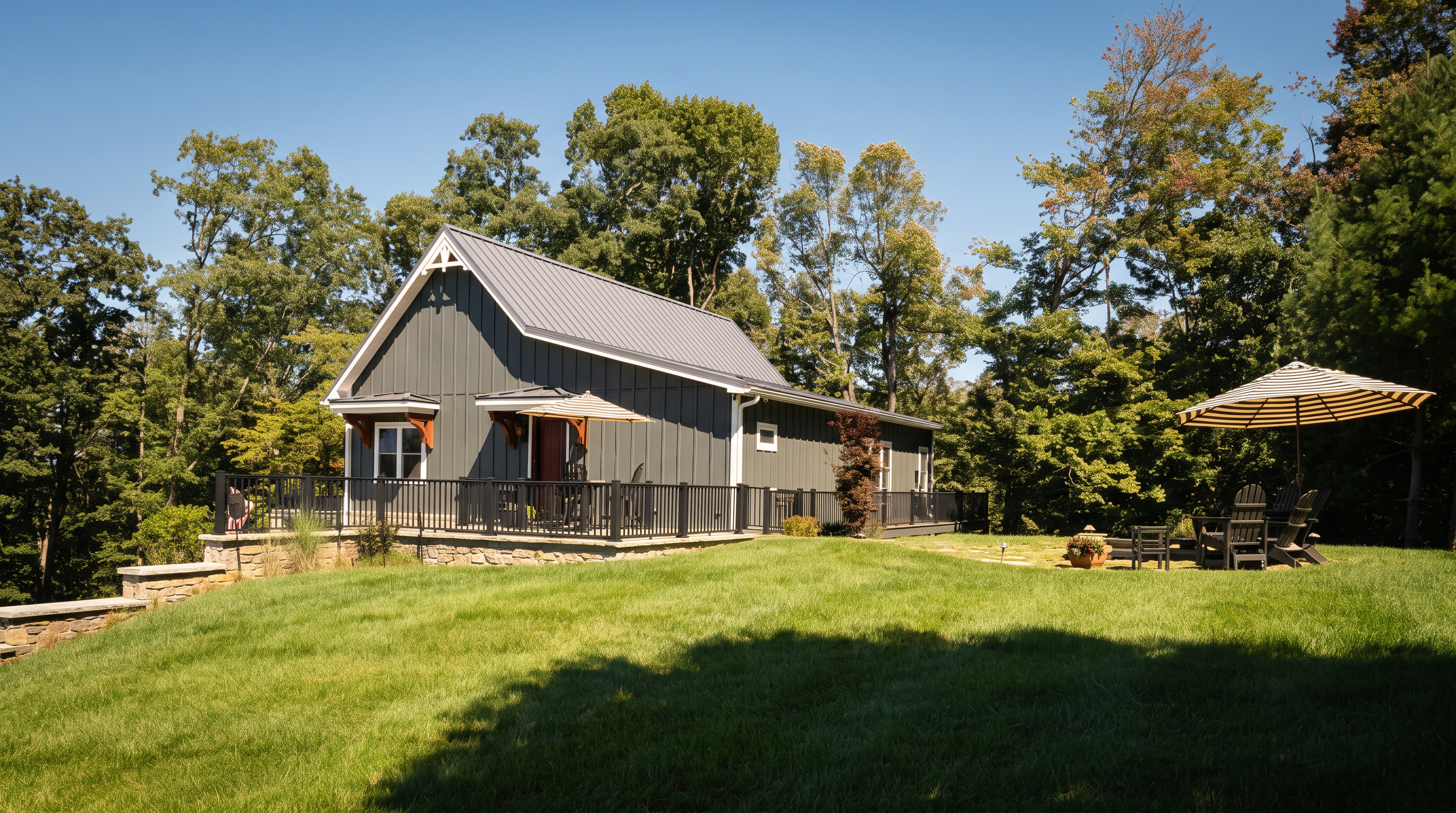 This exterior view captures the rear of a modern farmhouse-style home featuring dark vertical siding and a metal roof. A spacious stone patio with a black metal railing extends from the house, providing an ideal space for outdoor seating and relaxation. The property is set against a backdrop of lush green trees under a clear blue sky, with a well-manicured lawn in the foreground.