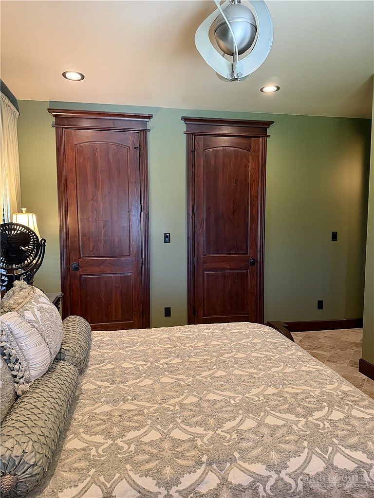 This bedroom features a neutral-toned patterned bedspread and two prominent, dark-stained wood doors with arched detailing set against sage green walls. A modern, spherical pendant light fixture hangs from the ceiling, while recessed lighting provides additional illumination. The room includes a glimpse of tiled flooring and a bedside lamp, creating a cozy and traditional atmosphere.