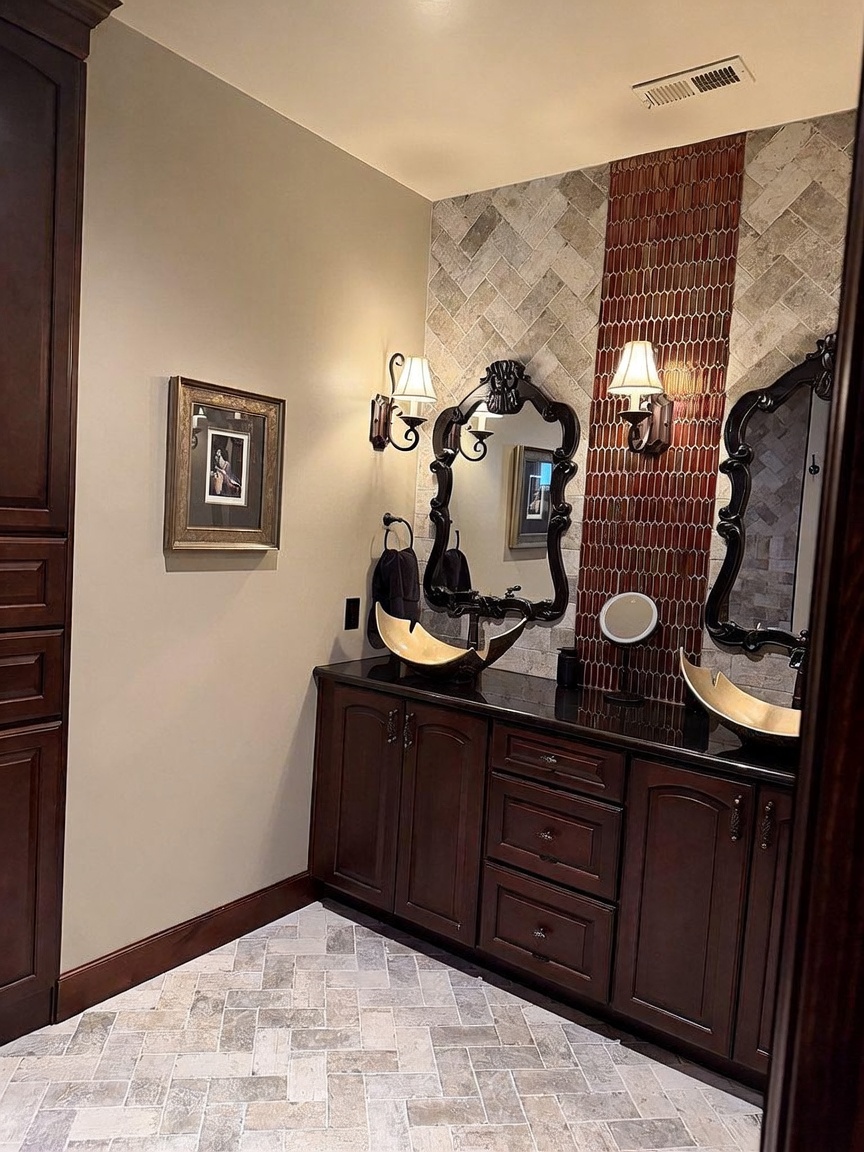 This elegant bathroom features a double vanity with dark wood cabinetry, a black countertop, and unique gold-toned vessel sinks. The wall behind the mirrors is accented with a vertical strip of narrow, reddish-brown tiles flanked by light-toned herringbone stone tile, which matches the flooring. Ornate black-framed mirrors and matching wall sconces add a sophisticated, traditional touch to the space.