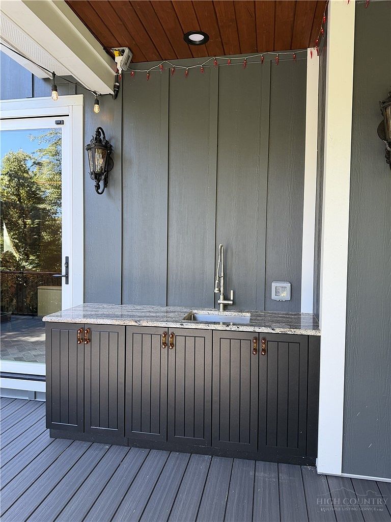 This outdoor living space features a built-in wet bar with dark, vertical-plank cabinetry and a patterned stone countertop. A stainless steel faucet and sink are integrated into the counter, set against a dark gray vertical-siding wall under a covered deck area. The scene is captured from a straight-on perspective, highlighting the functional outdoor kitchen setup on a composite deck.