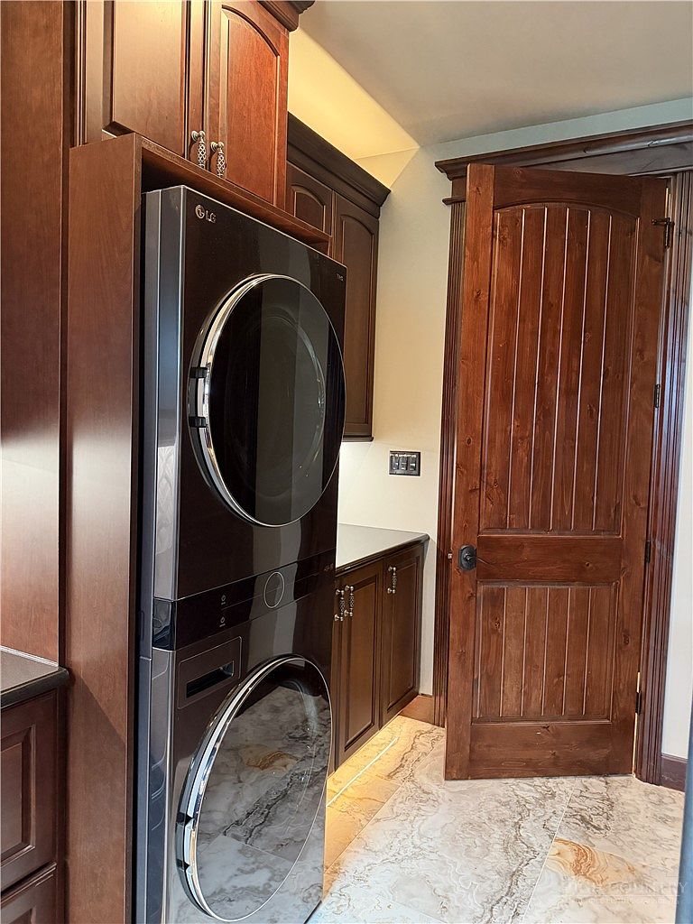This laundry room features a stacked LG washer and dryer unit integrated into custom dark wood cabinetry. The space includes a stone-topped counter and patterned tile flooring, creating a cohesive and functional design. A classic wooden door with a paneled design adds a traditional touch to the room's aesthetic.