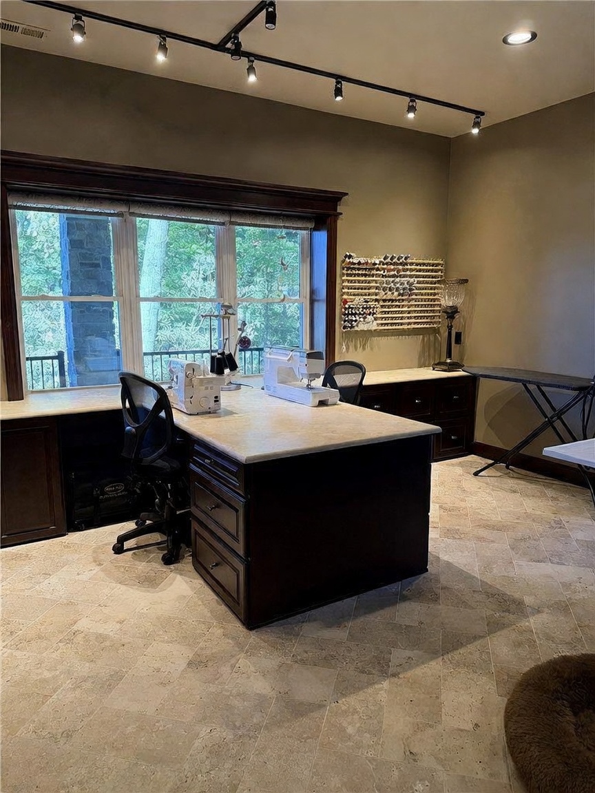 This dedicated craft or hobby room features a large central island with a stone-look countertop and dark cabinetry, providing ample workspace for sewing or creative projects. Large windows with dark wood trim offer a view of the outdoors, while a wall-mounted thread rack and track lighting enhance the functionality of the space. The room is finished with neutral-toned stone tile flooring, creating a clean and organized environment.