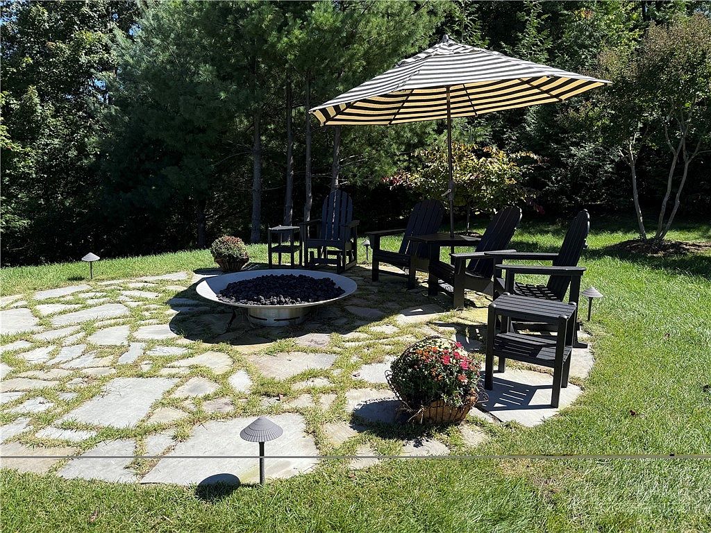 This outdoor living space features a circular flagstone patio set into a lush green lawn, centered around a modern, low-profile metal fire pit. A striped umbrella provides shade for a grouping of dark-toned Adirondack chairs and small side tables, creating a cozy seating area. The scene is framed by mature trees and landscaping, offering a peaceful and private backyard retreat.