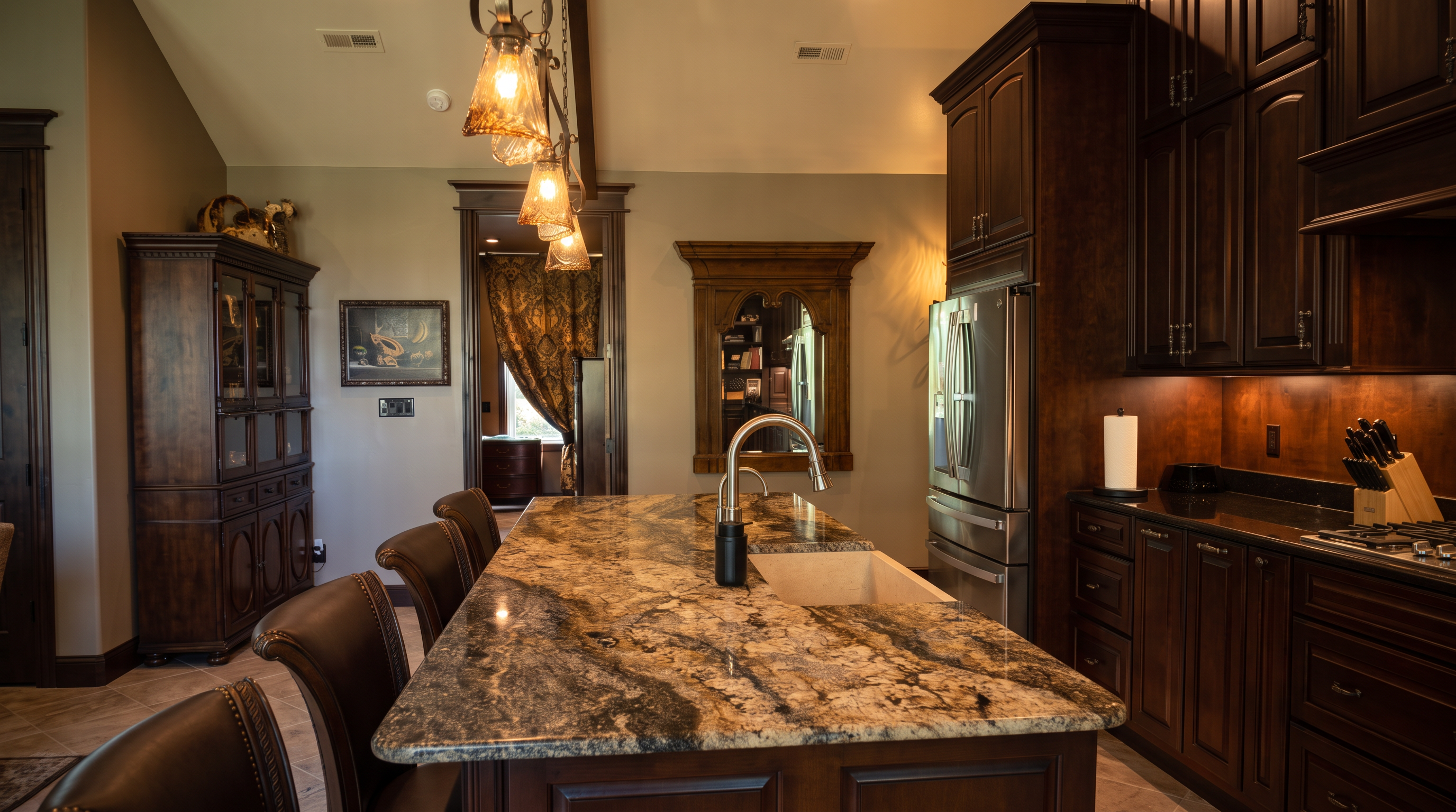 This kitchen features a large island with a prominent stone countertop and a farmhouse-style sink, serving as the central focal point. Dark wood cabinetry provides a rich contrast, while a decorative mirror framed in wood adds a touch of elegance to the wall behind the island. The perspective looks across the island toward a doorway leading into an adjacent room, creating a sense of depth and flow within the home.