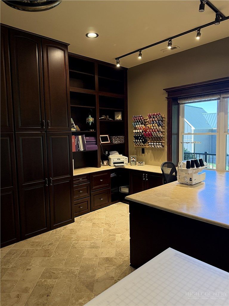 This dedicated craft or office space features extensive dark wood cabinetry and built-in shelving that provides ample storage. A large, L-shaped desk with a light-colored countertop offers a spacious workspace, complemented by a wall-mounted thread rack and a sewing machine on a central island. The room is illuminated by track lighting and natural light from a window, creating a functional and organized environment.