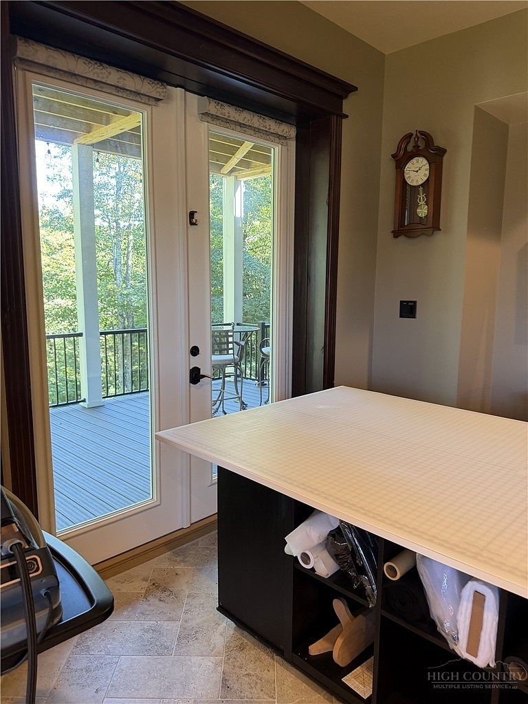 This interior space features a large, central work island with a grid-patterned cutting mat surface and open storage shelving underneath. A set of glass-paned double doors provides direct access to an outdoor deck, allowing for natural light and a view of the surrounding trees. A traditional wall-mounted clock hangs on the neutral-toned wall, adding a classic touch to the functional workspace.
