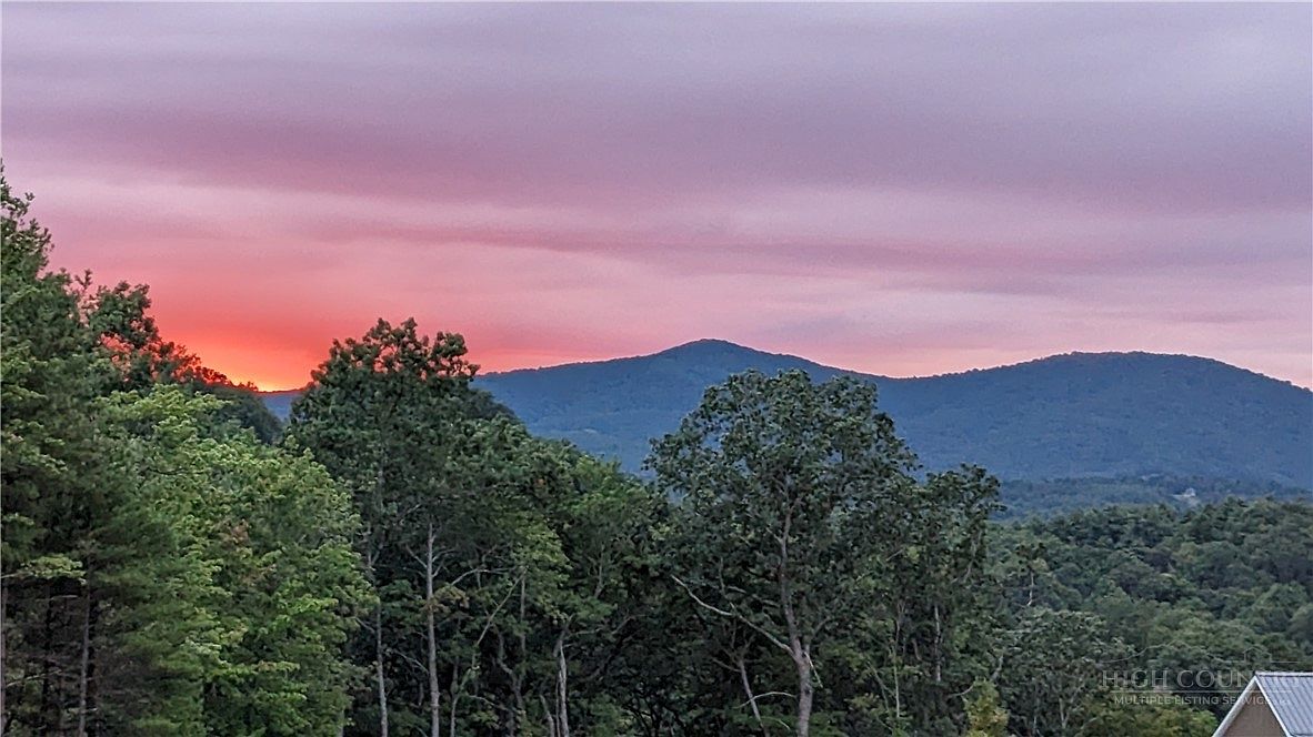 This image captures a scenic mountain landscape at sunset, showcasing rolling hills and dense forest foliage under a vibrant, pink-hued sky. The perspective is elevated, likely taken from a high vantage point or balcony, emphasizing the expansive natural views available from the property. The serene atmosphere highlights the peaceful, secluded nature of the location.